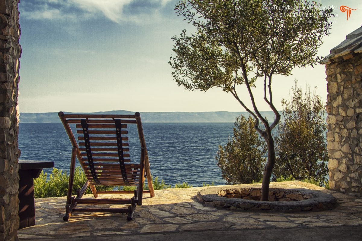 A wooden bedchair next to an olive tree and a traditional stone house with the sea view